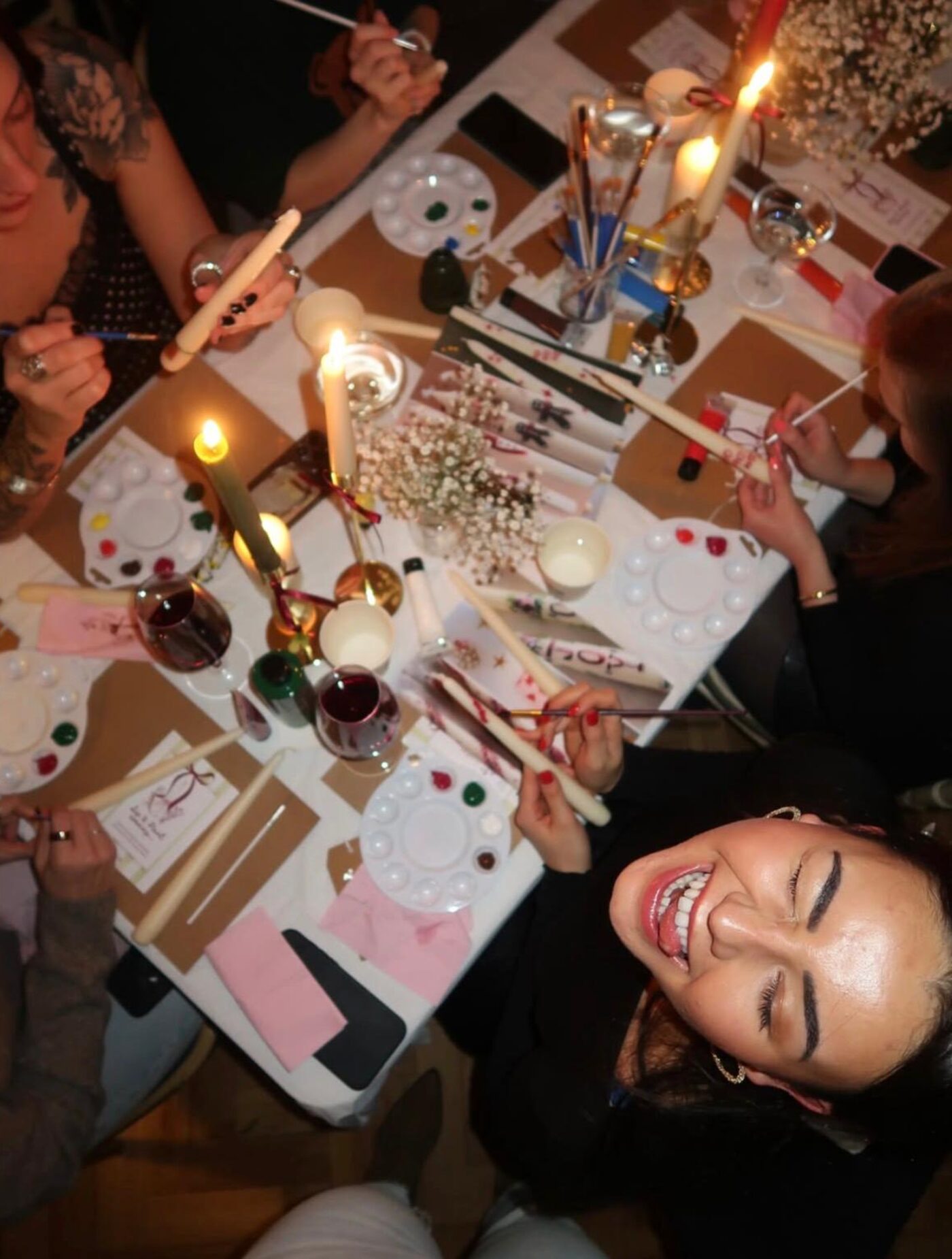 Looking down on a candlelit table of guests painting candles, one guest grinning up at the camera
