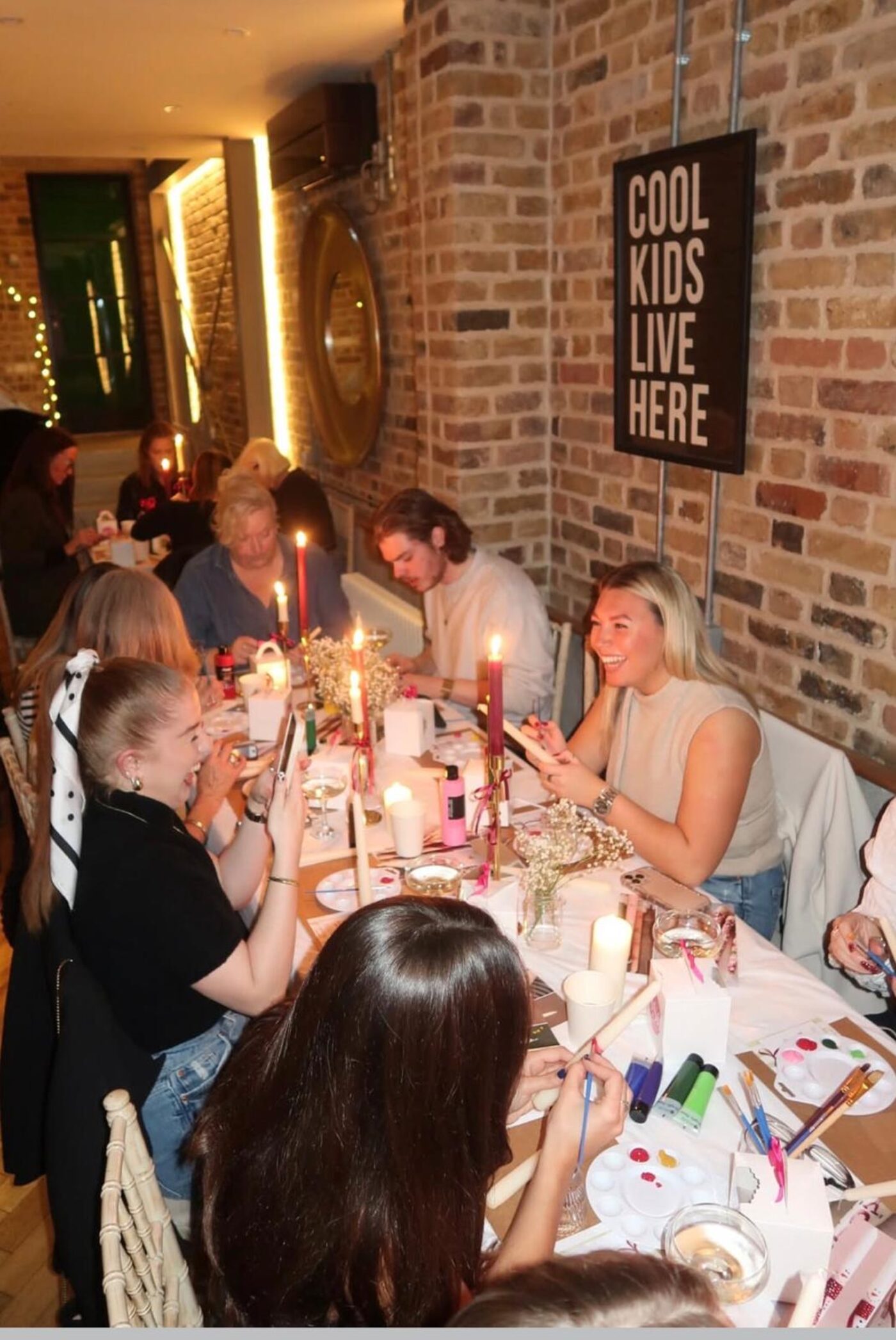 Long candlelit table of guests painting and laughing under a Cool Kids Live Here sign