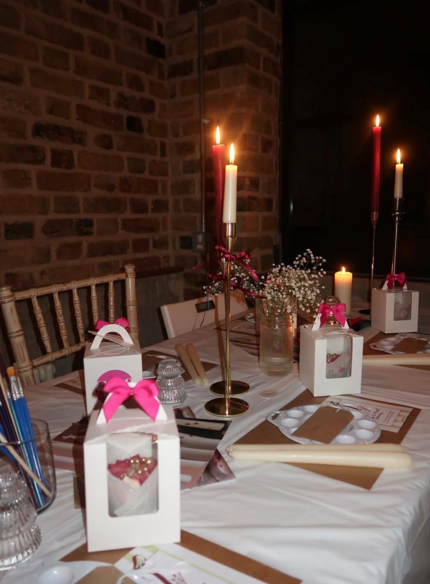 Table set for a candle painting workshop with tall candles, gift boxes tied with pink bows and paint palettes
